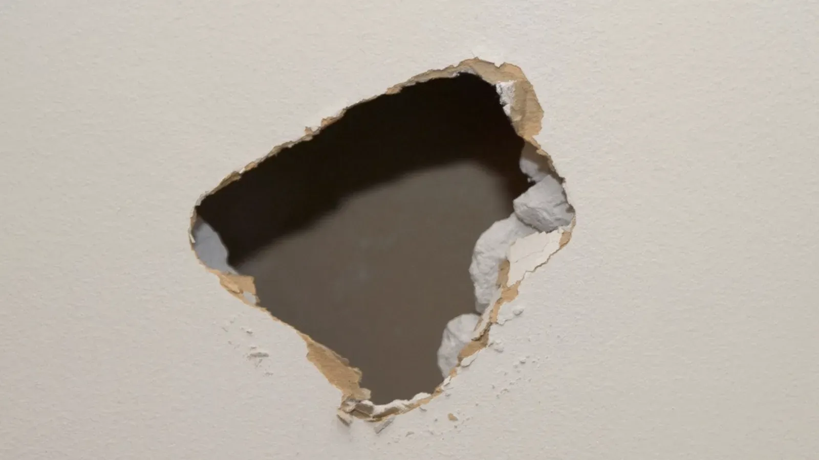 Jagged impact hole broken through a painted interior drywall wall, torn paper facing and crumbling gypsum around the opening exposing the dark wall cavity behind