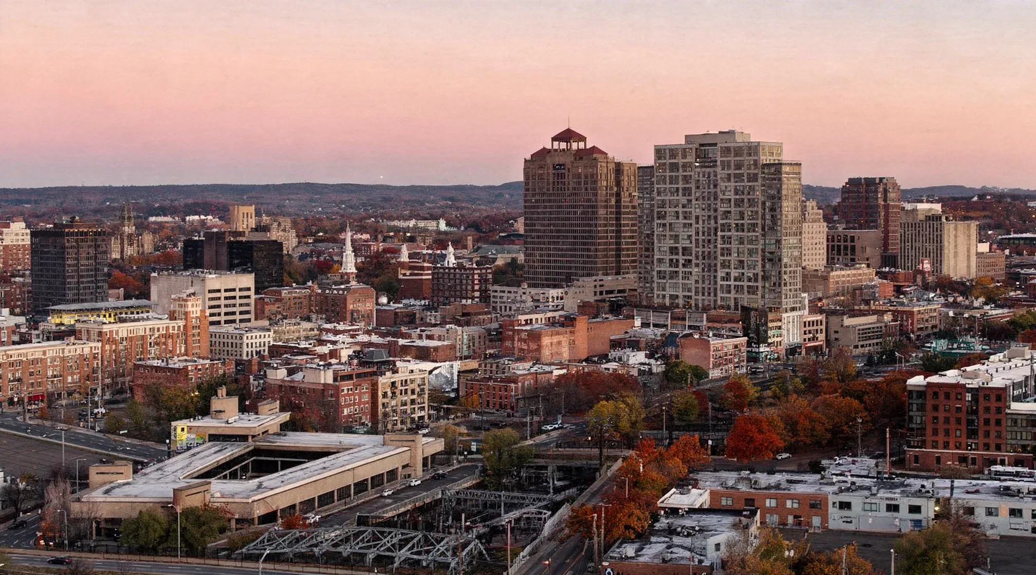 Downtown New Haven skyline at dusk, the city's high-rise office and residential towers rising above brick low-rise blocks and autumn trees